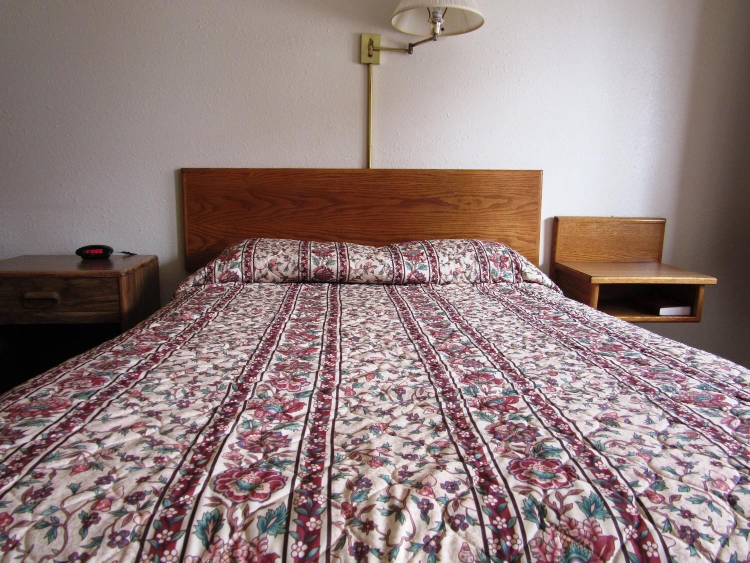 A bed with a floral-patterned bedspread, a wooden headboard, a bedside table with a lamp, and a nightstand with a clock.