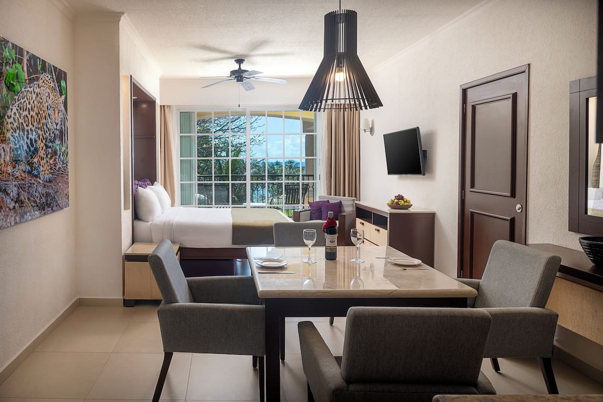 Una habitación de hotel con una cama, un área de comedor con mesa y sillas, un televisor montado en la pared y un balcón con vistas al exterior.
