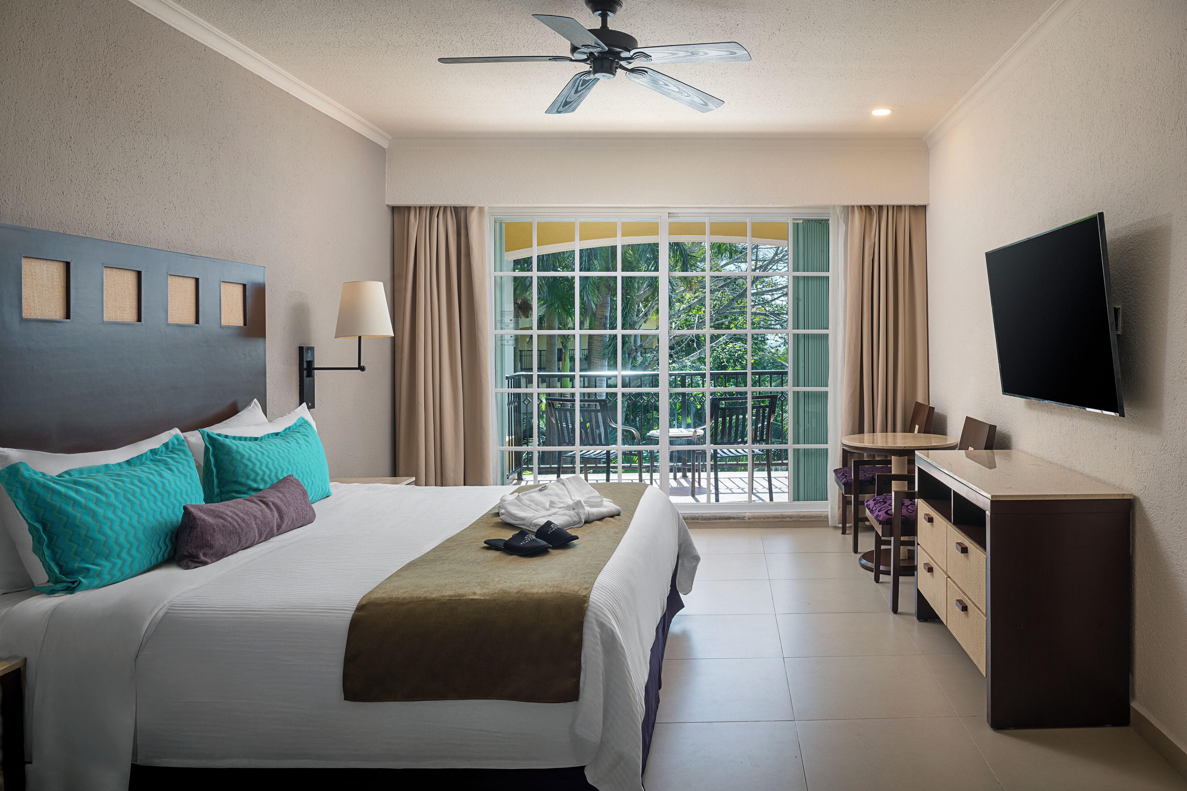 A hotel room with a bed, a TV, a desk, and a balcony with a view of trees.