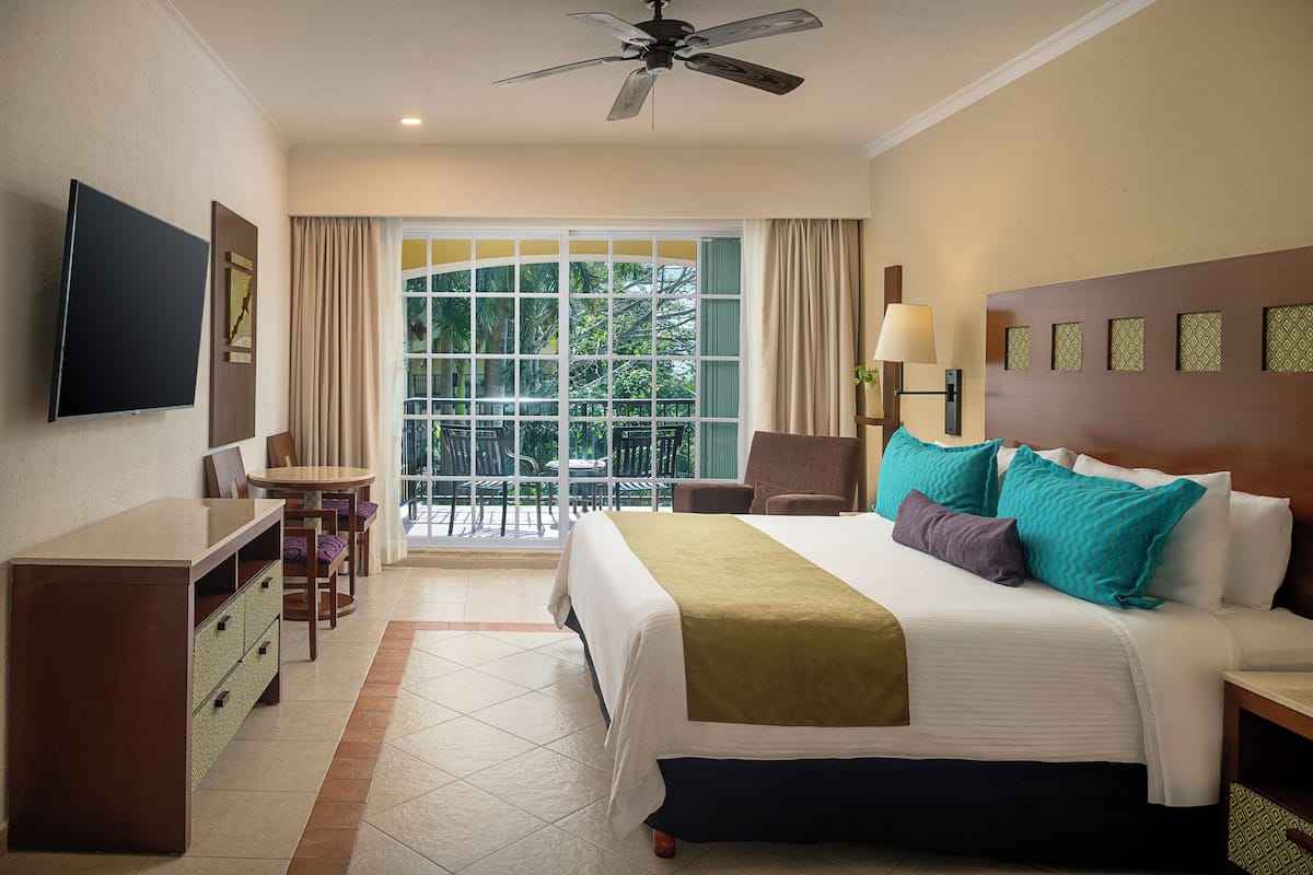 Habitación de hotel con cama, escritorio, silla, televisión y balcón con vista a árboles.