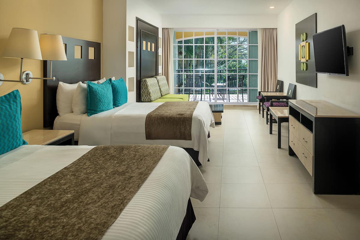 Habitación de hotel con dos camas, un escritorio, un televisor y un balcón con vistas al follaje.