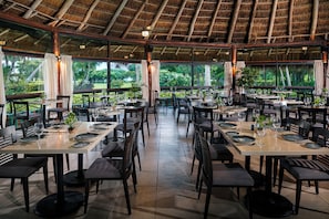 An indoor dining area with tables set for guests, featuring a thatched roof and large windows overlooking a lush garden.
