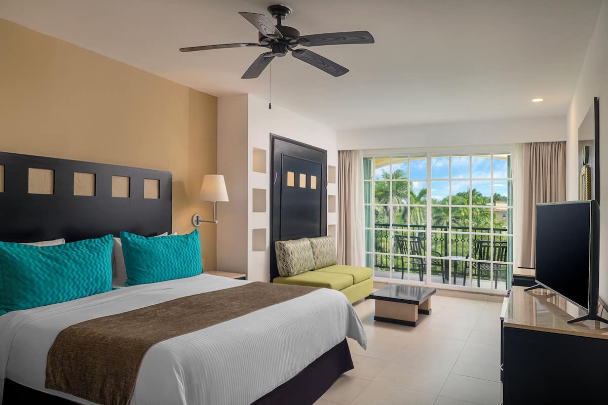 Habitación de hotel con cama, sofá, televisión, ventilador de techo y ventana con vistas a zonas verdes.