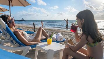 A beach scene with a person reclining on a sunbed, another person sitting on a chair, and a table with a drink and towels.