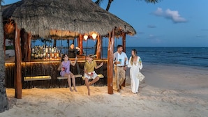 A beach bar with a thatched roof, wooden poles, and a bar counter with bottles. - Hotel Marina El Cid Spa & Beach Resort All Inclusive (Puerto Morelos)
