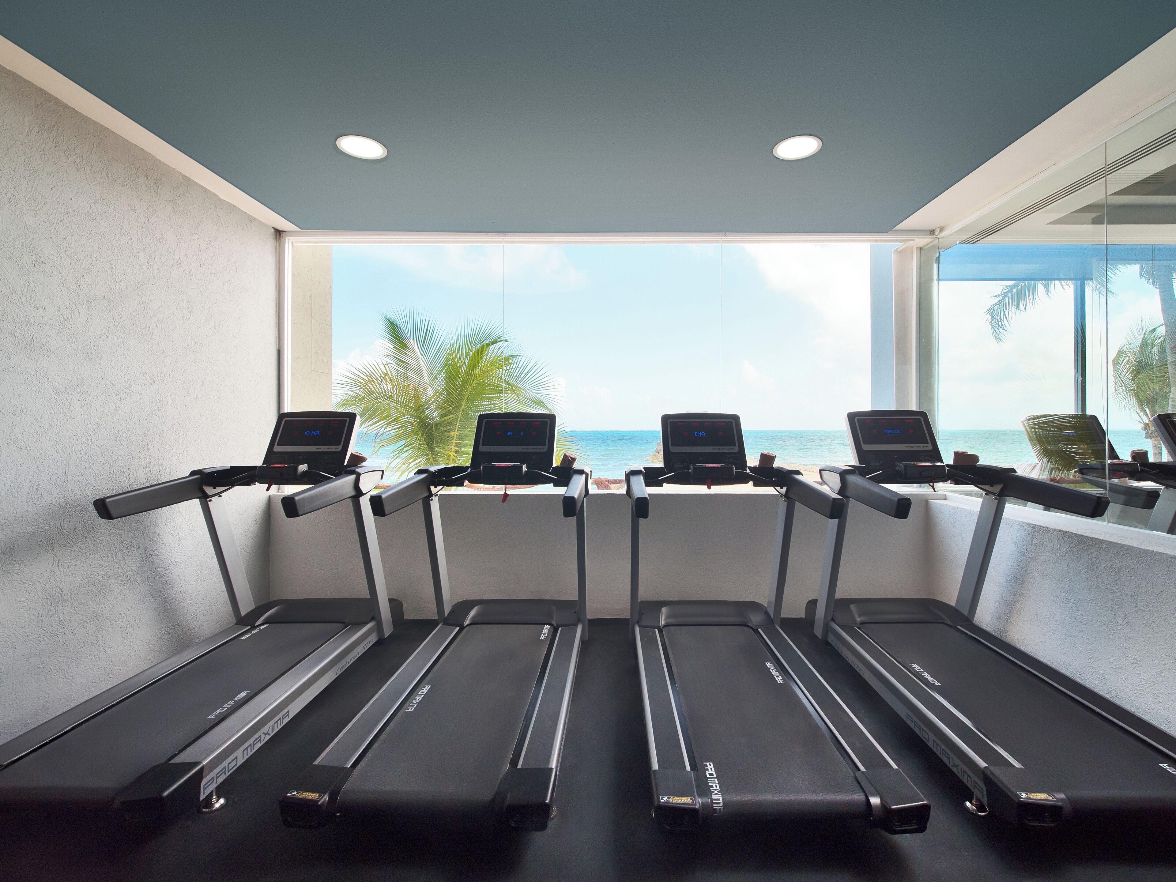 Four ProMaxima treadmills aligned in a row with a view of the ocean through large windows.