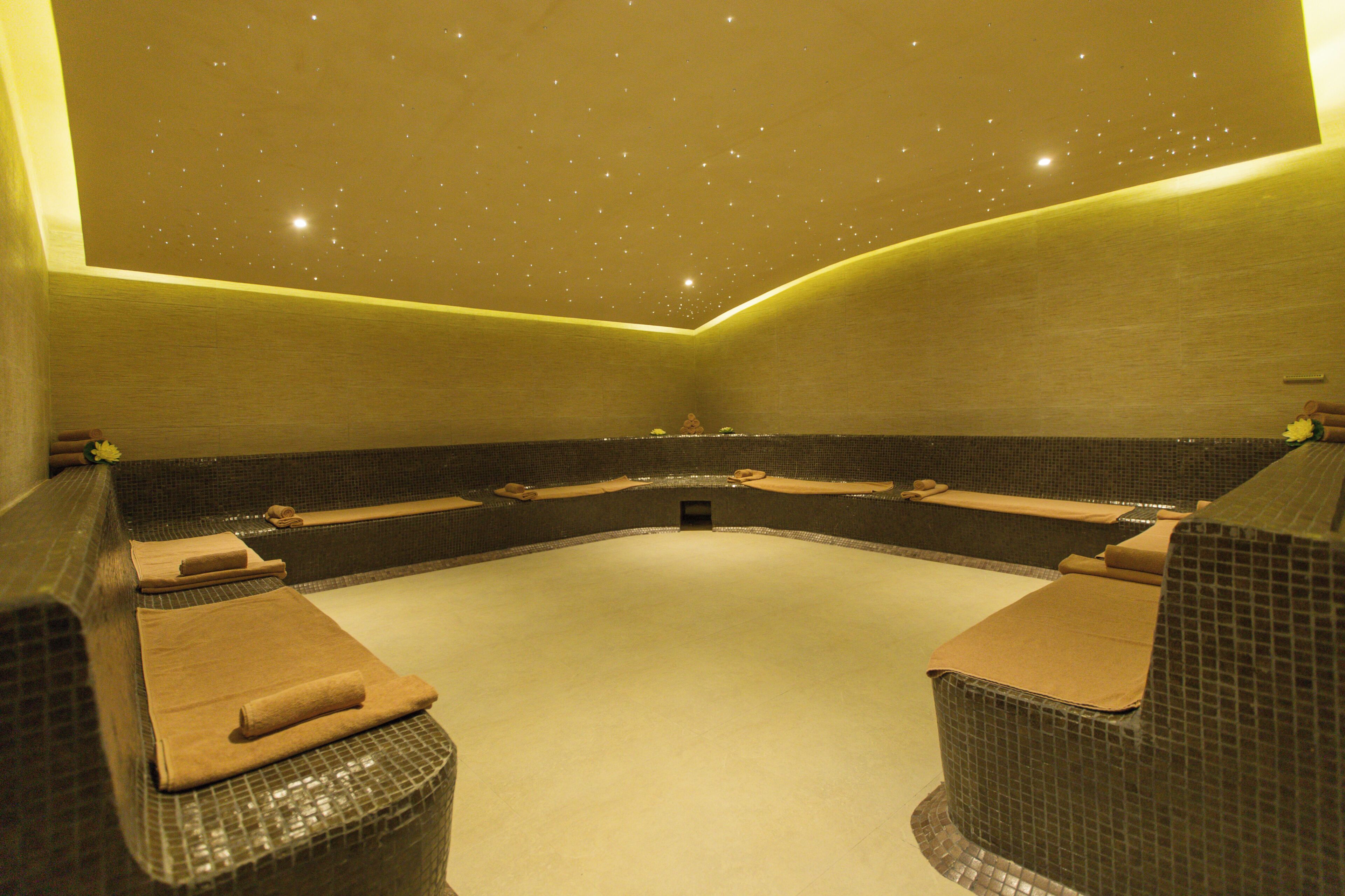 Un cuarto de spa con una bañera grande y curvada, dos zonas de descanso con toallas, y un techo con una decoración luminosa que simula estrellas.