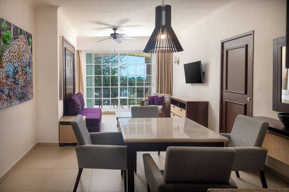 Habitación de hotel moderna con comedor, ventilador de techo, ventana grande con vistas y televisor de pantalla plana montado en la pared.