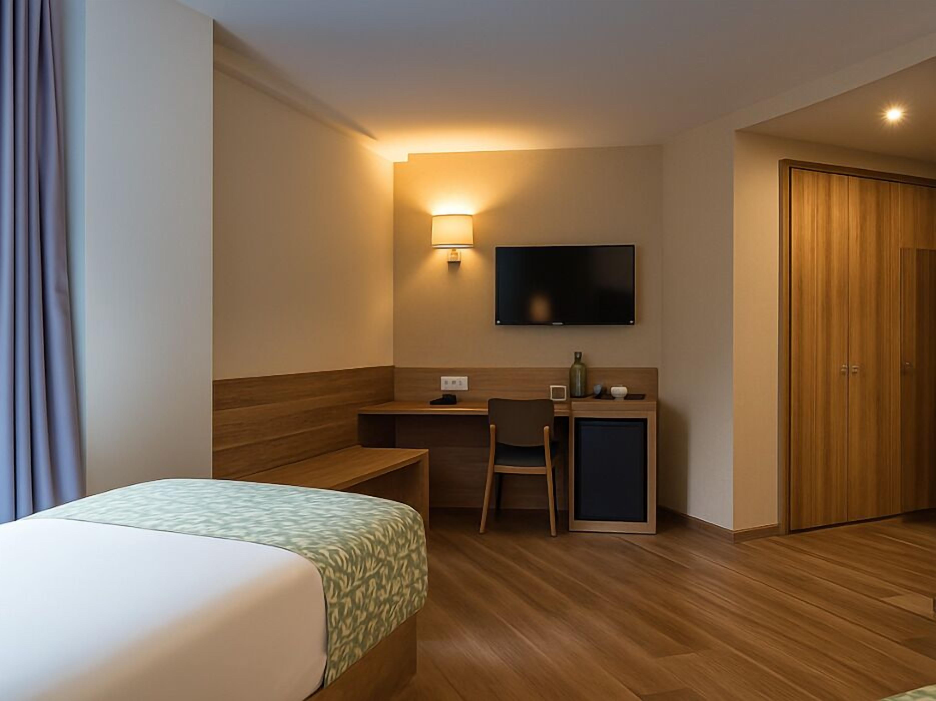 A hotel room with a bed, a desk with a chair, a television, and a wooden wardrobe.