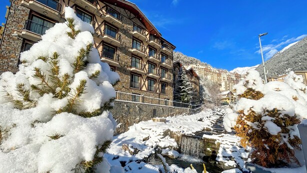A multi-story hotel with stone facade and balconies, a snow-covered landscape, and a small waterfall.