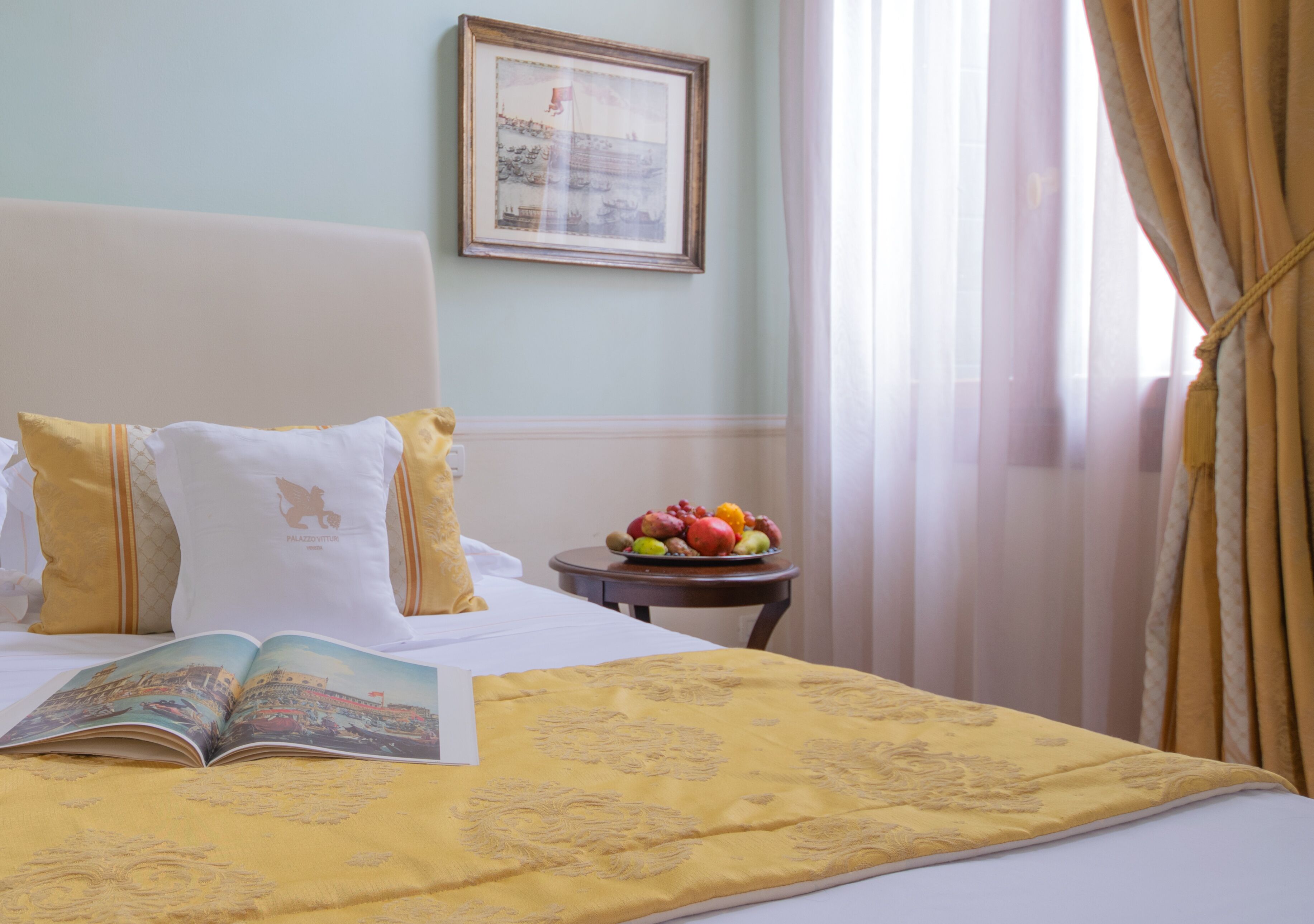 Un lit bien fait, recouvert d’une couverture jaune, un bol de fruits posé sur une petite table et un tableau encadré accroché au mur.