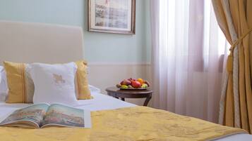 Un lit bien fait, recouvert d’une couverture jaune, un bol de fruits posé sur une petite table et un tableau encadré accroché au mur.