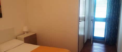 Habitación de hotel con cama, mesita de noche, lámpara y vista a un pasillo con puerta y ventana.