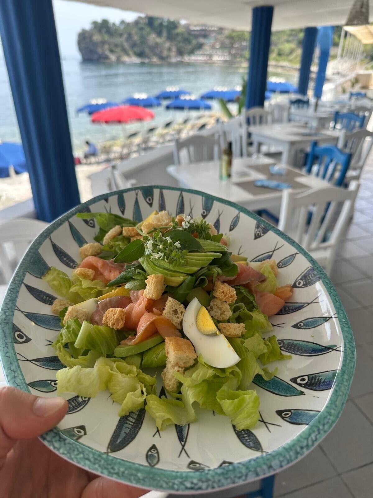 A bowl of salad with various ingredients, held in a hand, with a view of a beach and umbrellas in the background.