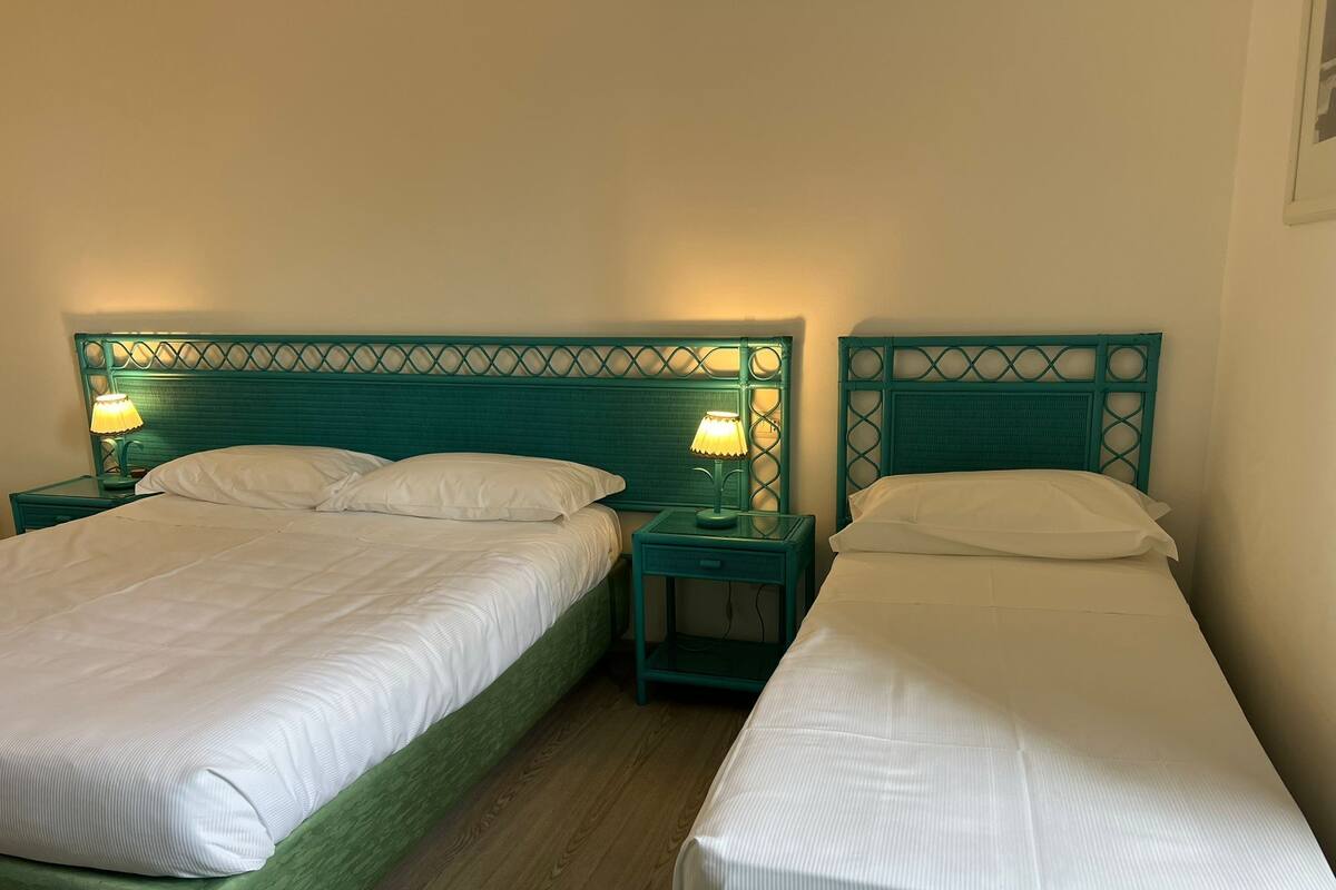 A hotel room with two single beds, each with a green headboard and white linens. There are bedside tables with lamps on each side of the beds.