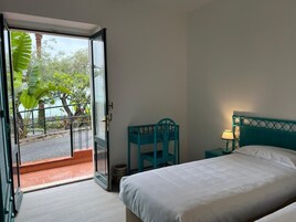 Une chambre avec un lit, une table de chevet, un fauteuil turquoise et une vue sur un balcon agrémenté d’une balustrade et de plantes, accessible par une porte ouverte.