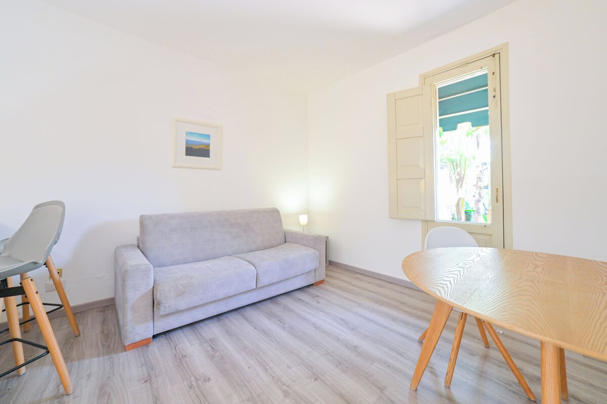 Ein Zimmer mit einer hellgrauen Couch, einem Holzesstisch und einem Fenster mit Blick auf Grünflächen.