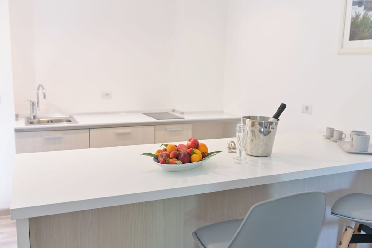 Cozinha moderna com bancada branca, uma tigela de frutas, uma chaleira e uma pia.