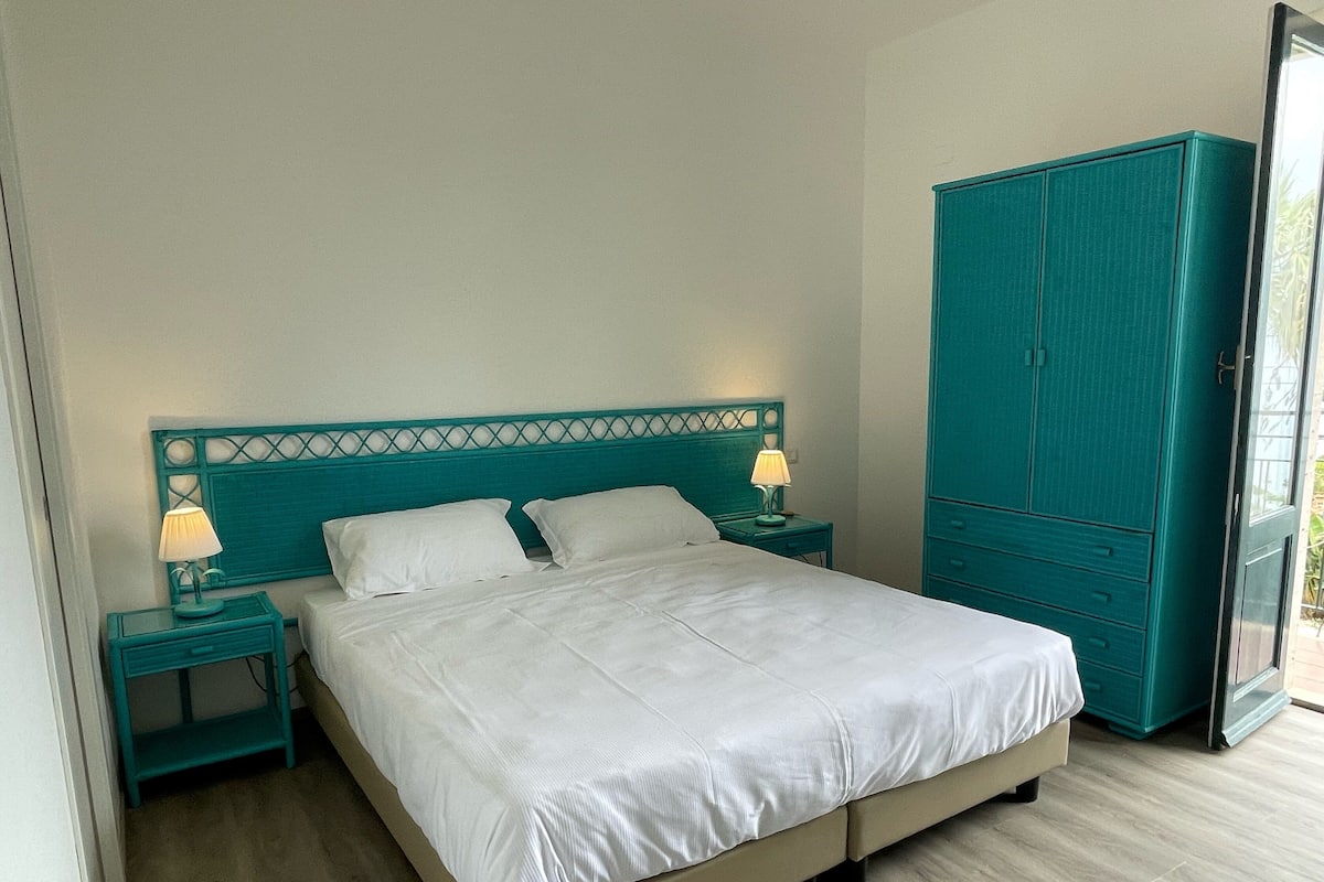 A bedroom with a bed, two bedside tables with lamps, a wardrobe, and a view of the outdoors.