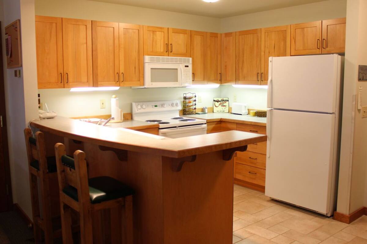 A kitchen with wooden cabinets, a white microwave, a stove, a refrigerator, and a bar counter with chairs.