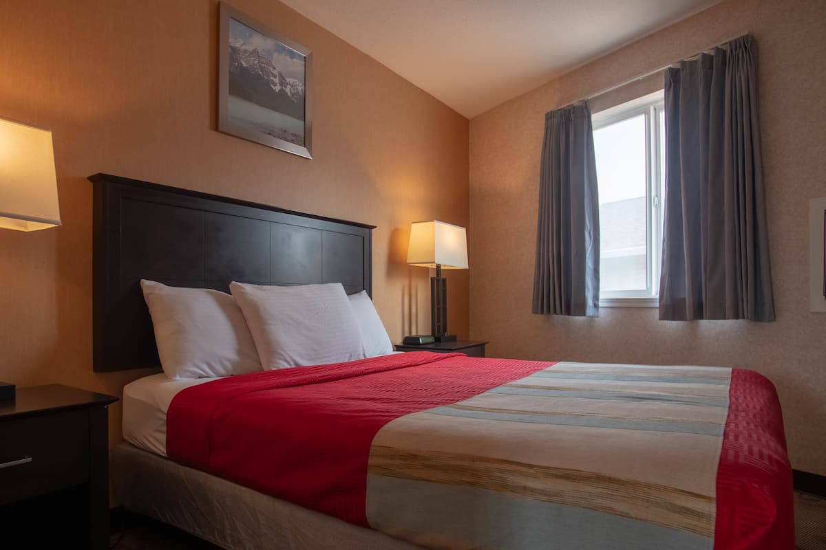 Ein Hotelzimmer mit einem Bett, einem Nachttisch, einer Lampe, einem Fenster mit Vorhängen und einem gerahmten Bild an der Wand.