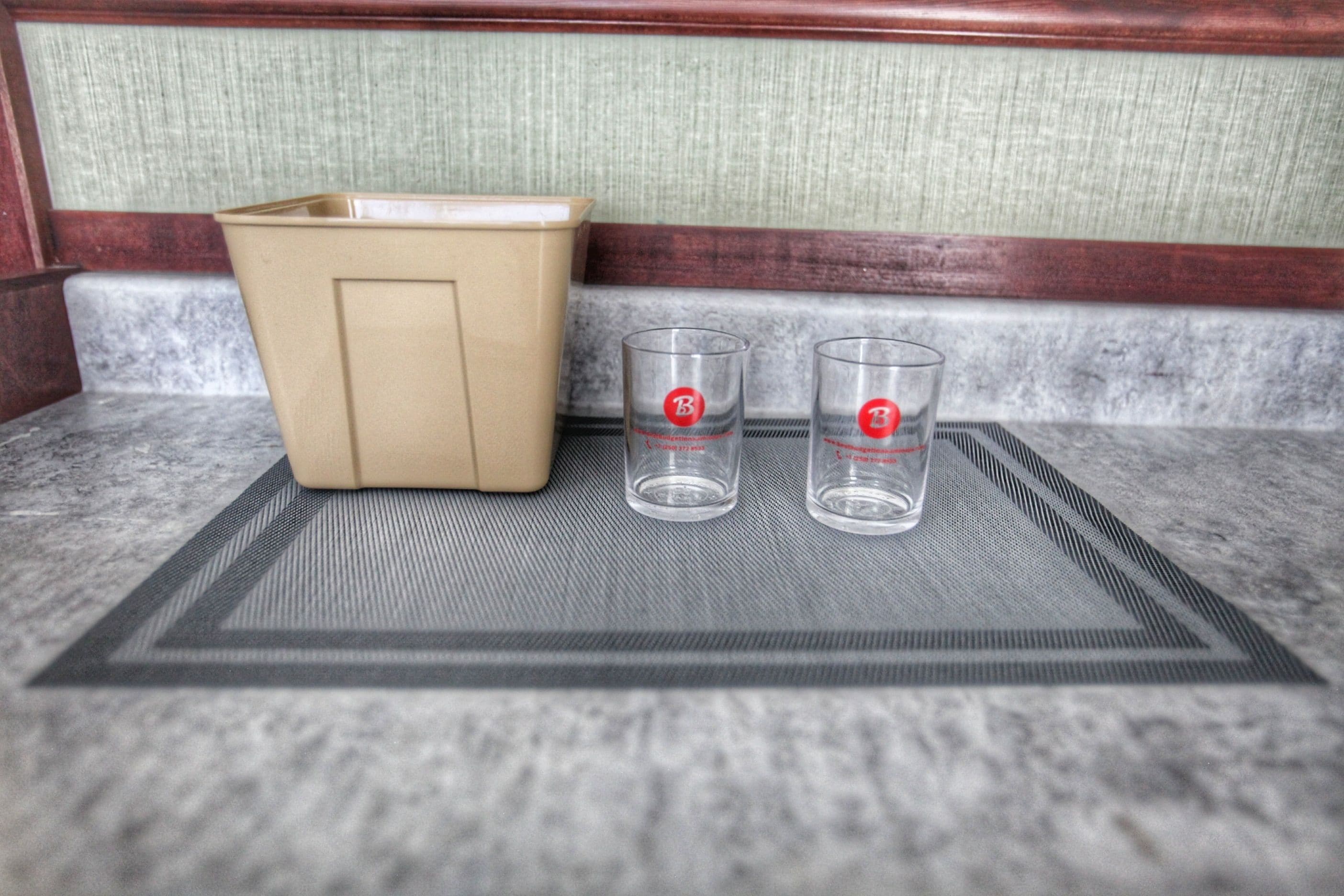 Una esquina de una habitación con un cubo de basura color beige y dos vasos transparentes con logotipos rojos sobre un tapete texturizado.
