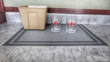 Una esquina de una habitación con un cubo de basura color beige y dos vasos transparentes con logotipos rojos sobre un tapete texturizado.