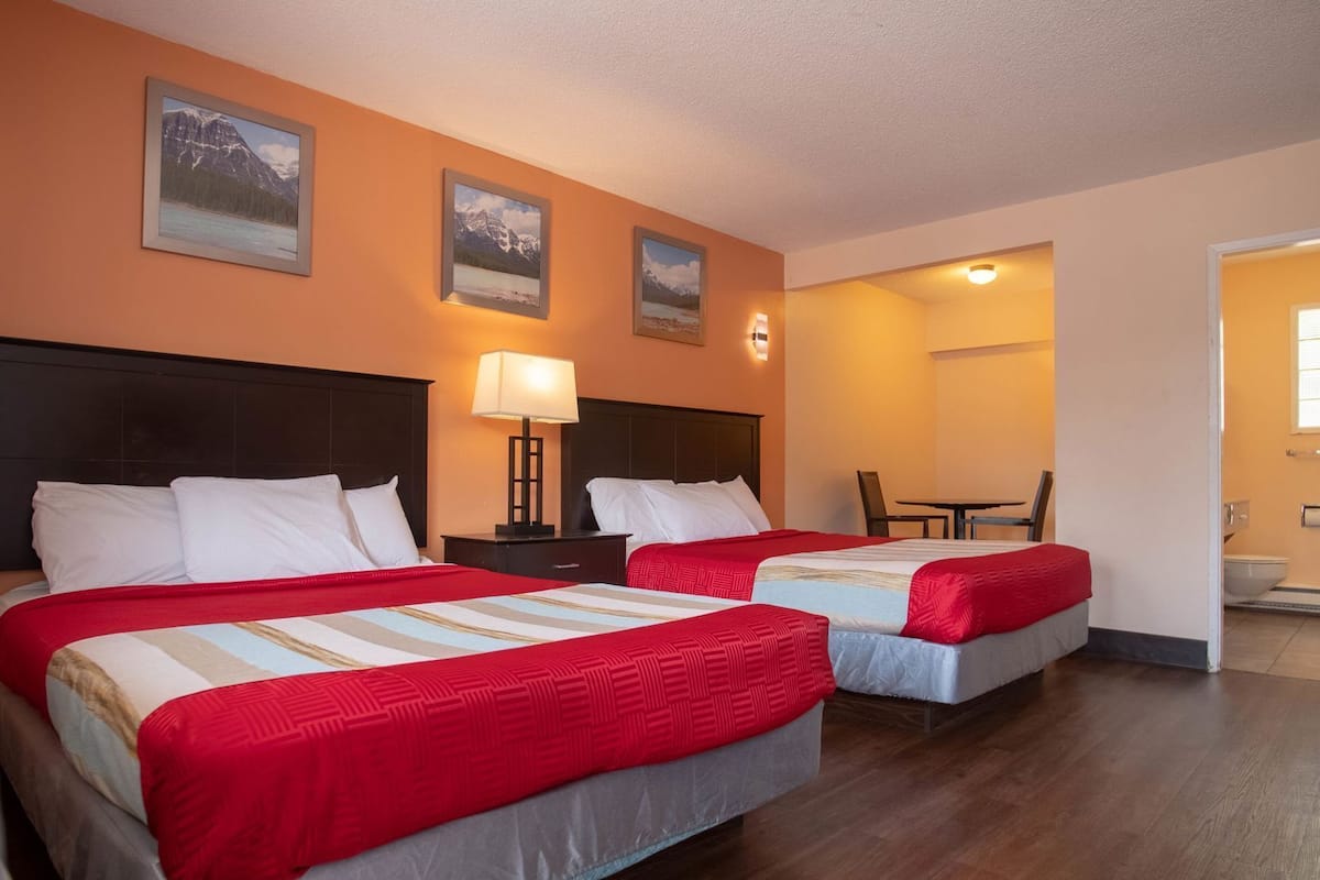 Ein Hotelzimmer mit zwei Betten, einem Nachttisch mit Lampe, einem Esstisch und Blick ins Bad.