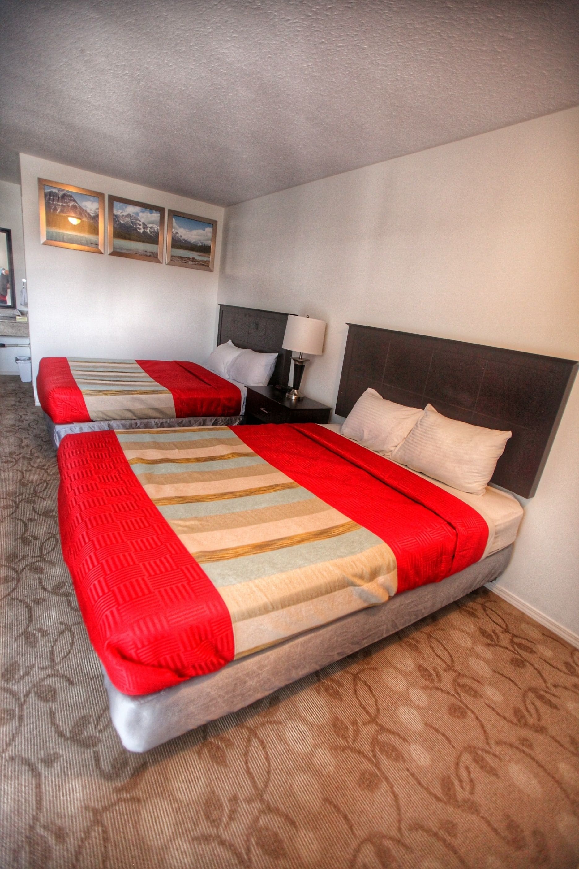 A hotel room with two beds, each with a red and beige striped bedspread, a nightstand with a lamp, and a wall-mounted picture above the beds.