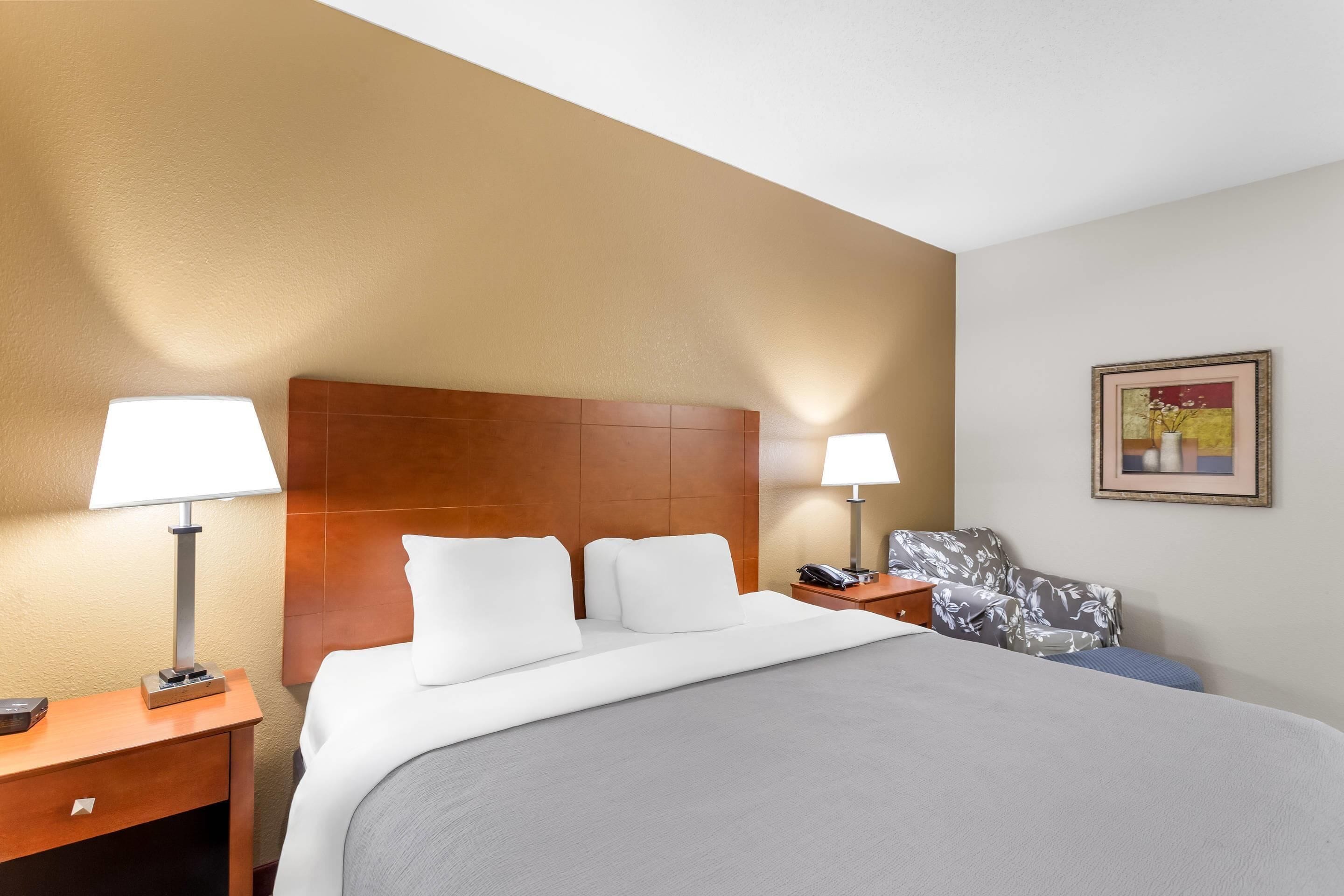 A hotel room with a large bed, two bedside tables with lamps, a wooden headboard, a floral-patterned chair, and a framed picture on the wall.