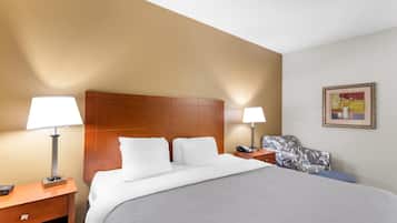 A hotel room with a large bed, two bedside tables with lamps, a wooden headboard, a floral-patterned chair, and a framed picture on the wall.