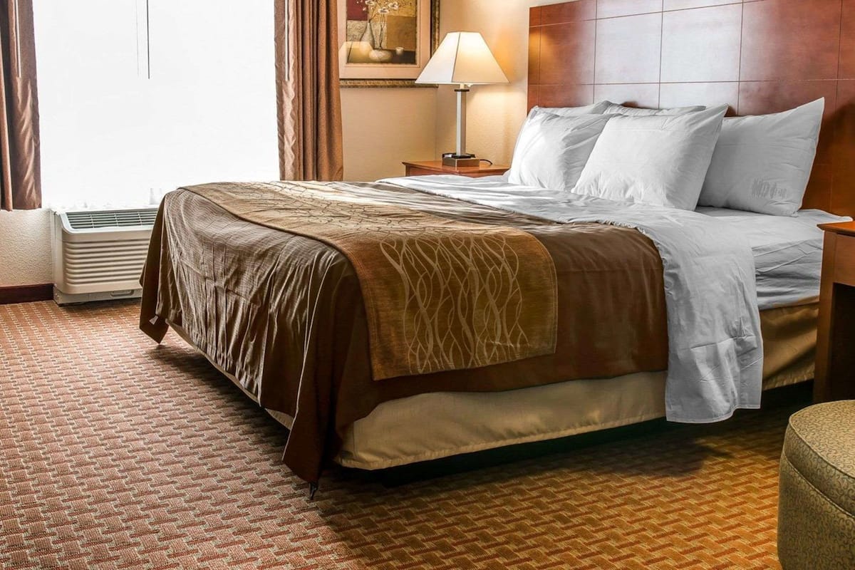 A hotel room with a large bed, a nightstand, a lamp, a painting on the wall, and a window with curtains.