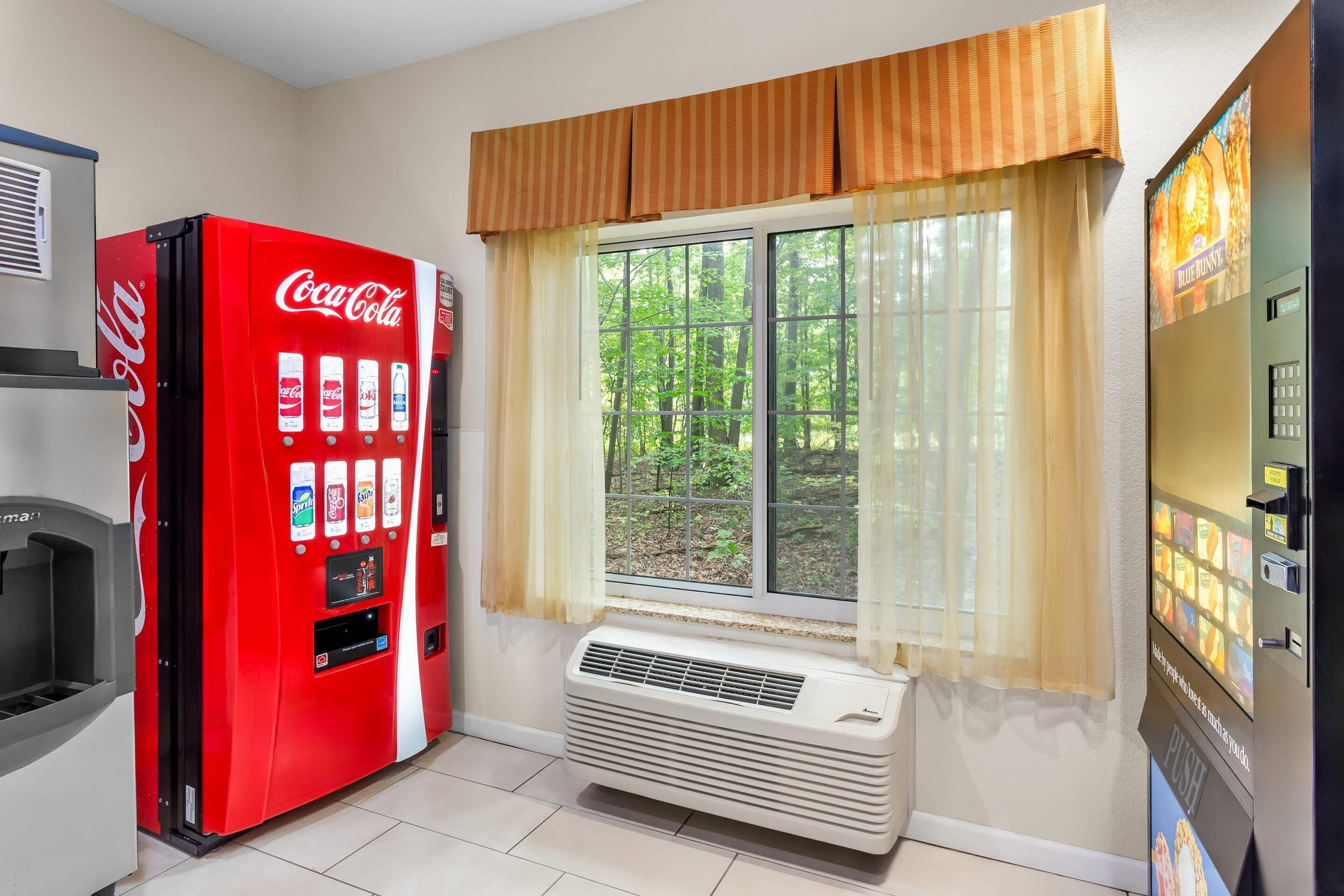 A room with a red Coca-Cola vending machine, a window with a view of greenery, and a wall-mounted air conditioner.