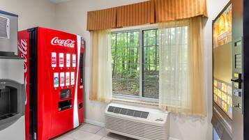 A room with a red Coca-Cola vending machine, a window with a view of greenery, and a wall-mounted air conditioner.
