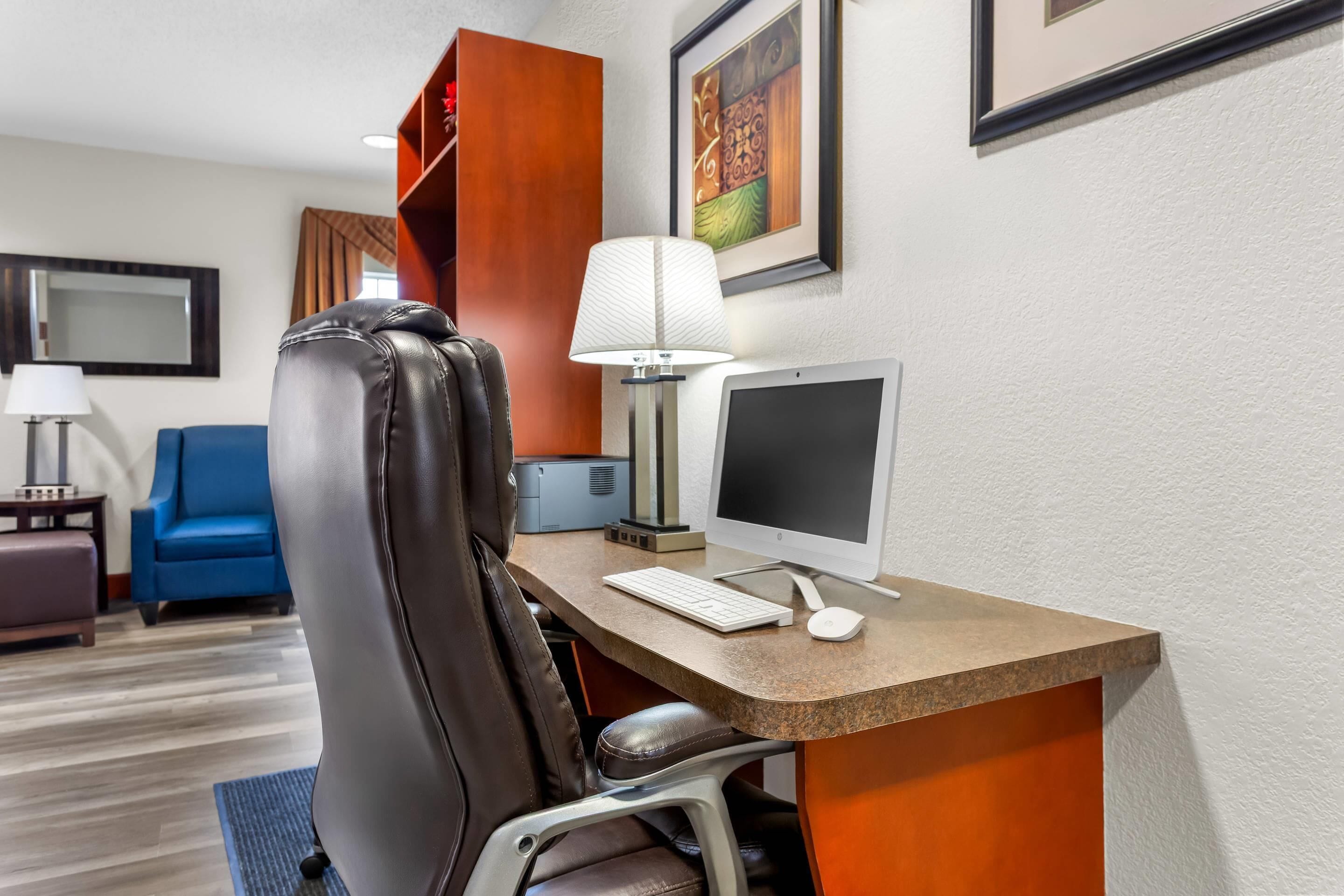 A hotel room with a desk, a computer, a chair, a lamp, a blue sofa, a small table with a lamp, and a framed picture on the wall.