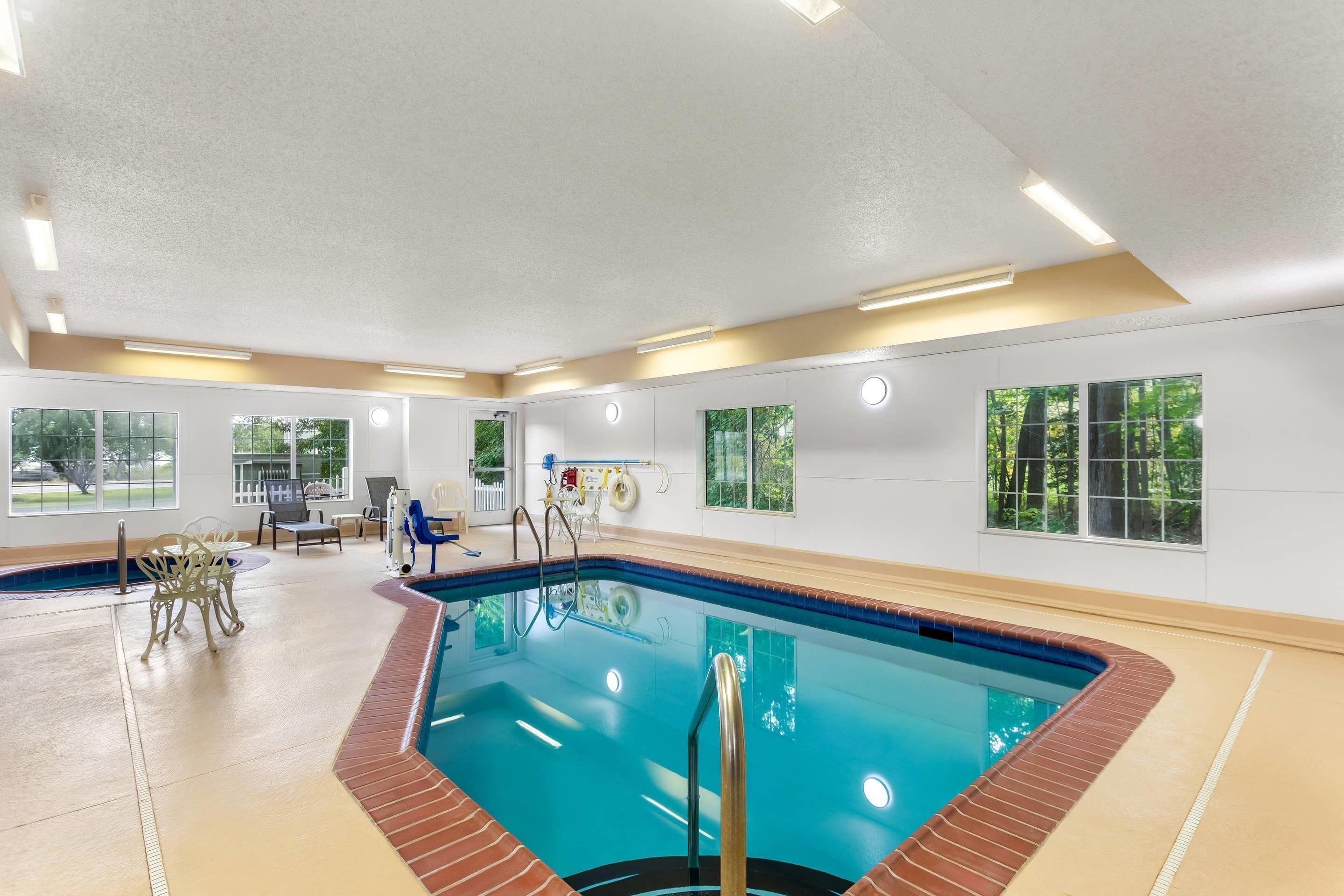 An indoor swimming pool with clear blue water, surrounded by a beige floor and walls, with windows offering views of greenery outside.