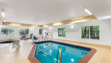 An indoor swimming pool with clear blue water, surrounded by a beige floor and walls, with windows offering views of greenery outside.