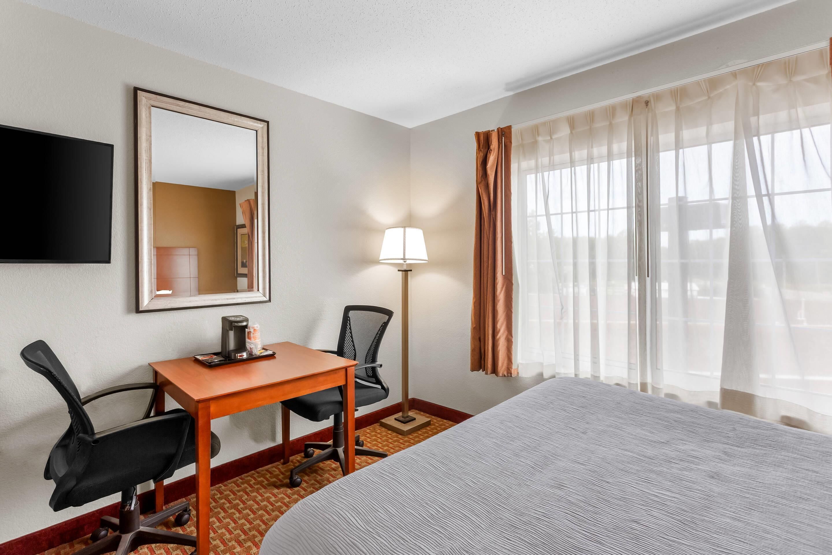 Une chambre d’hôtel comprenant un lit, un bureau avec une chaise, une lampe, un téléviseur et une fenêtre avec des rideaux.