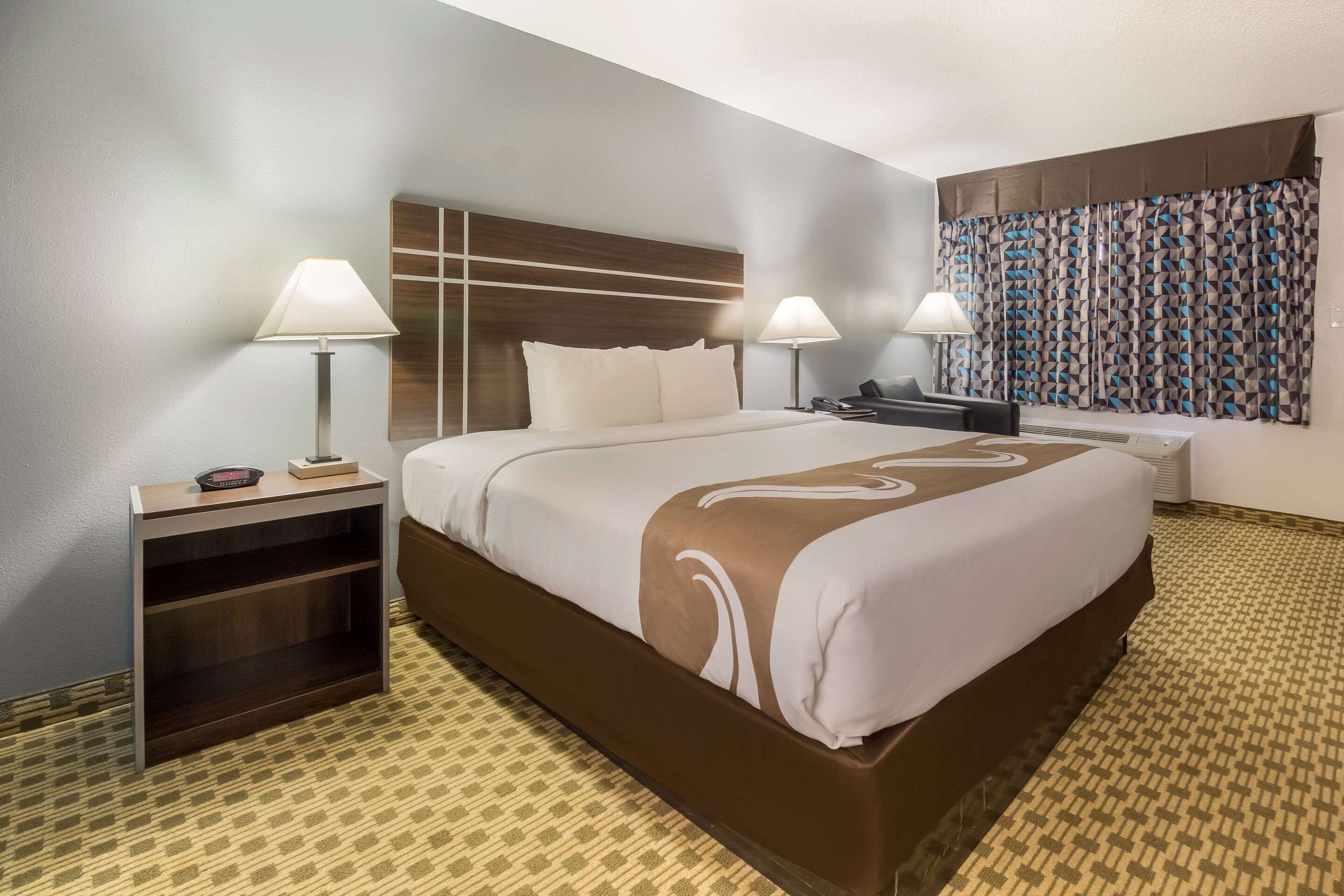 Una habitación de hotel con una cama grande, dos mesitas de noche con lámparas, un escritorio y una alfombra estampada.