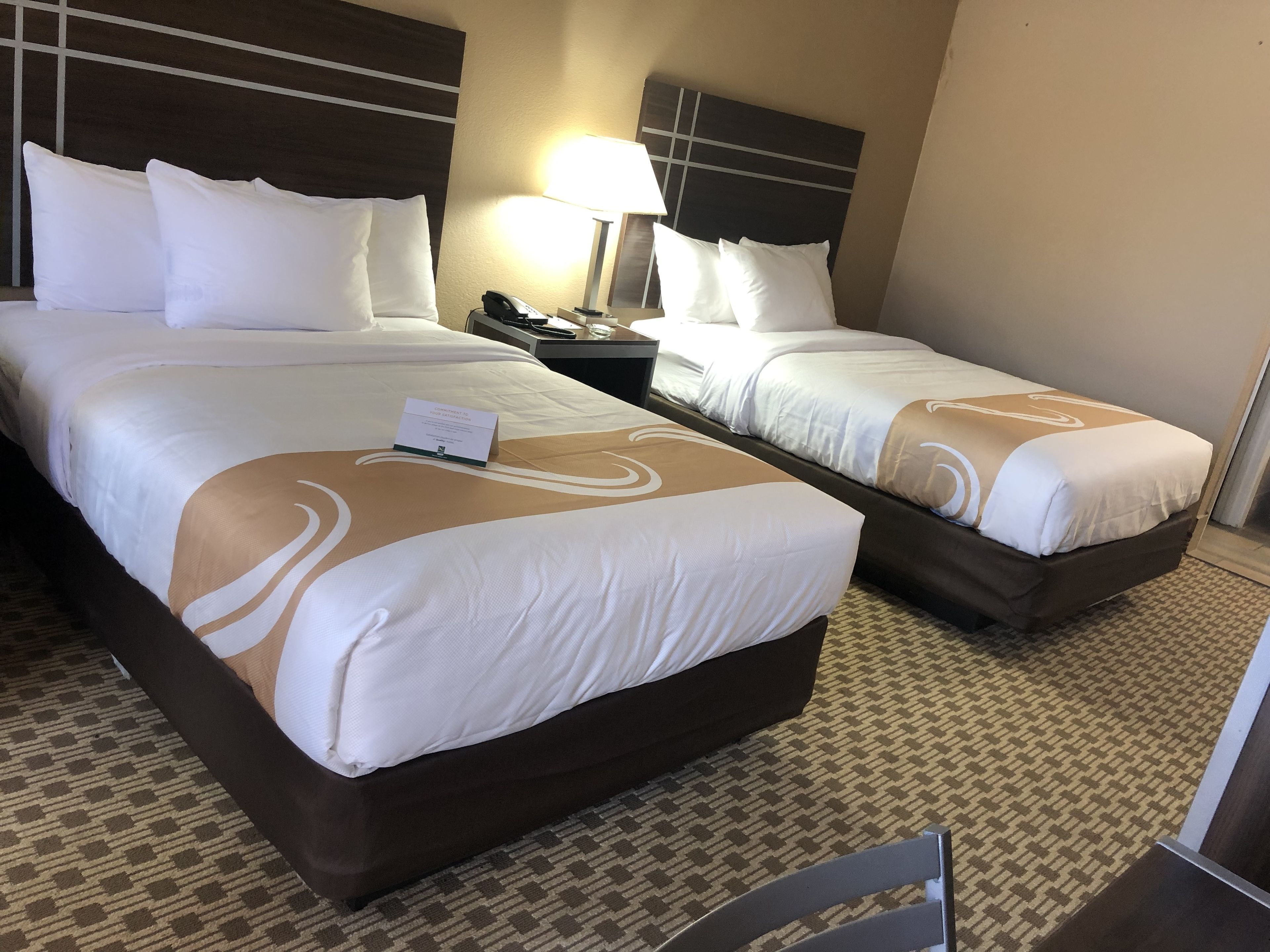 A hotel room with two beds, each with white bedding and a brown headboard. There is a nightstand with a lamp and a phone between the beds. The room has a patterned carpet and a small table with a chair.