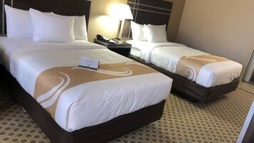 A hotel room with two beds, each with white bedding and a brown headboard. There is a nightstand with a lamp and a phone between the beds. The room has a patterned carpet and a small table with a chair.