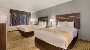 A hotel room with two beds, a large window with patterned curtains, a bedside table with a lamp, and a carpeted floor. - Quality Inn Madisonville (Madisonville)