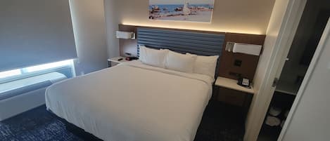Quarto de hotel com uma cama grande, cabeceira, mesa de cabeceira com abajur, quadro na parede e janela com persianas.