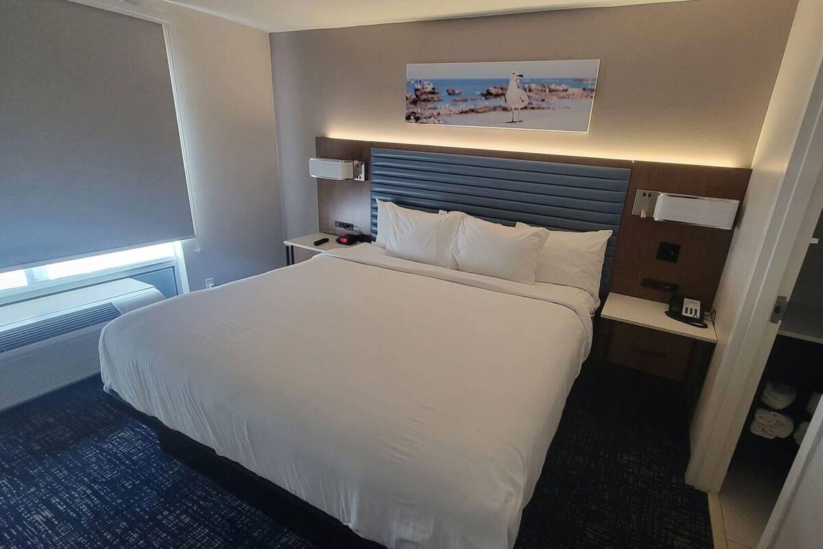 Une chambre d’hôtel avec un grand lit, des tables de chevet, une petite table, une lampe fixée au mur, un tableau et une fenêtre avec des stores.
