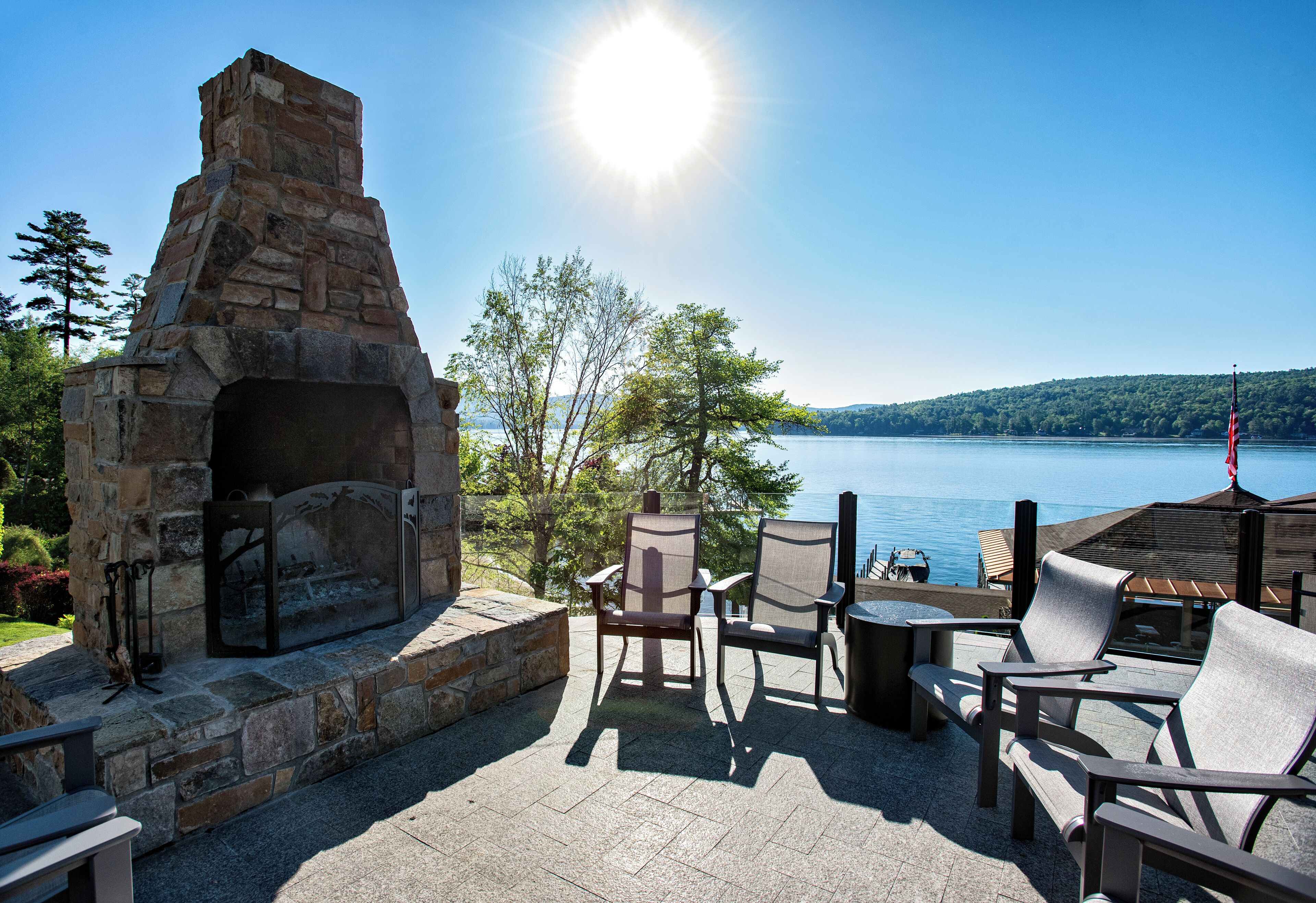 An outdoor patio with stone fireplace, seating, and a view of a lake.