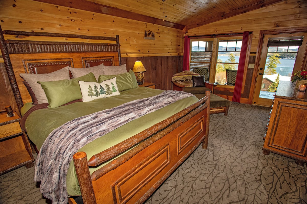 A wooden bedroom with a large bed, a nightstand, a chair, and a view of the outdoors.