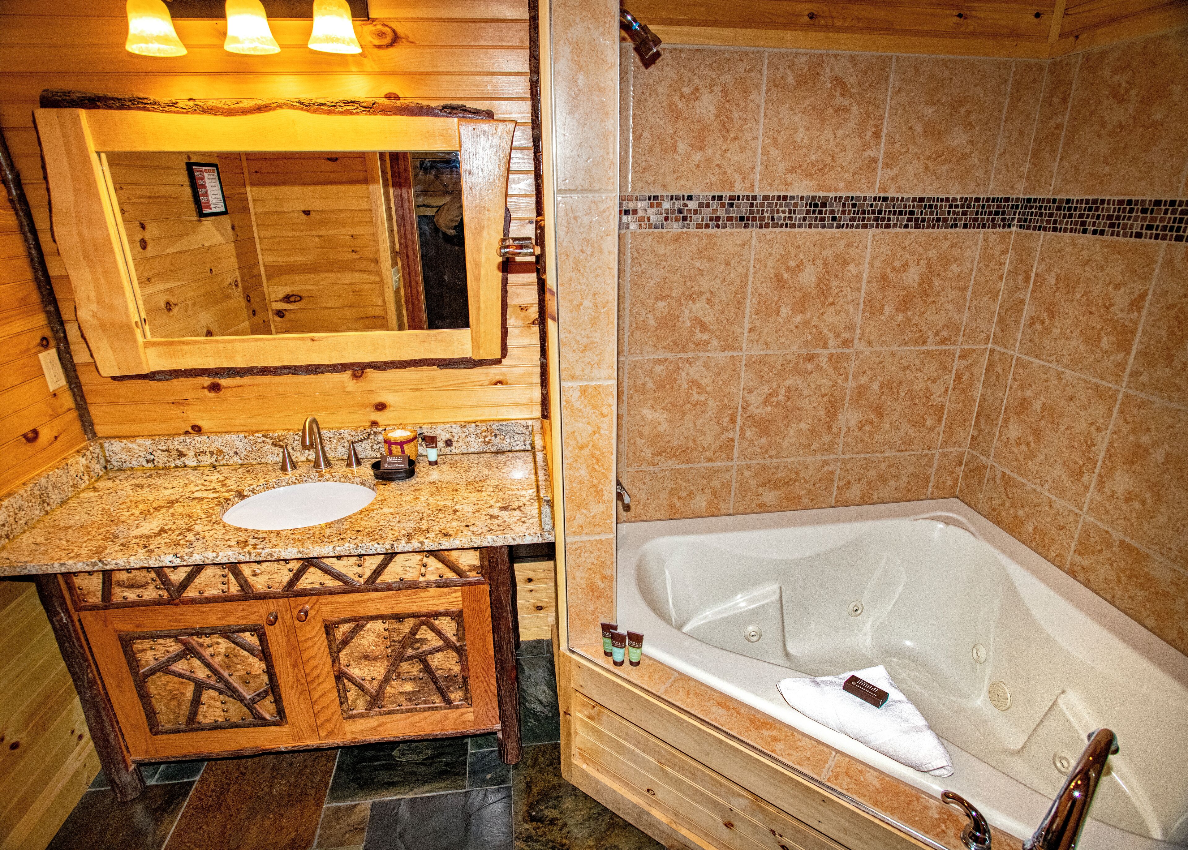 Un baño de cabaña de madera con lavamanos de granito, tocador de madera, un espejo grande y una bañera.