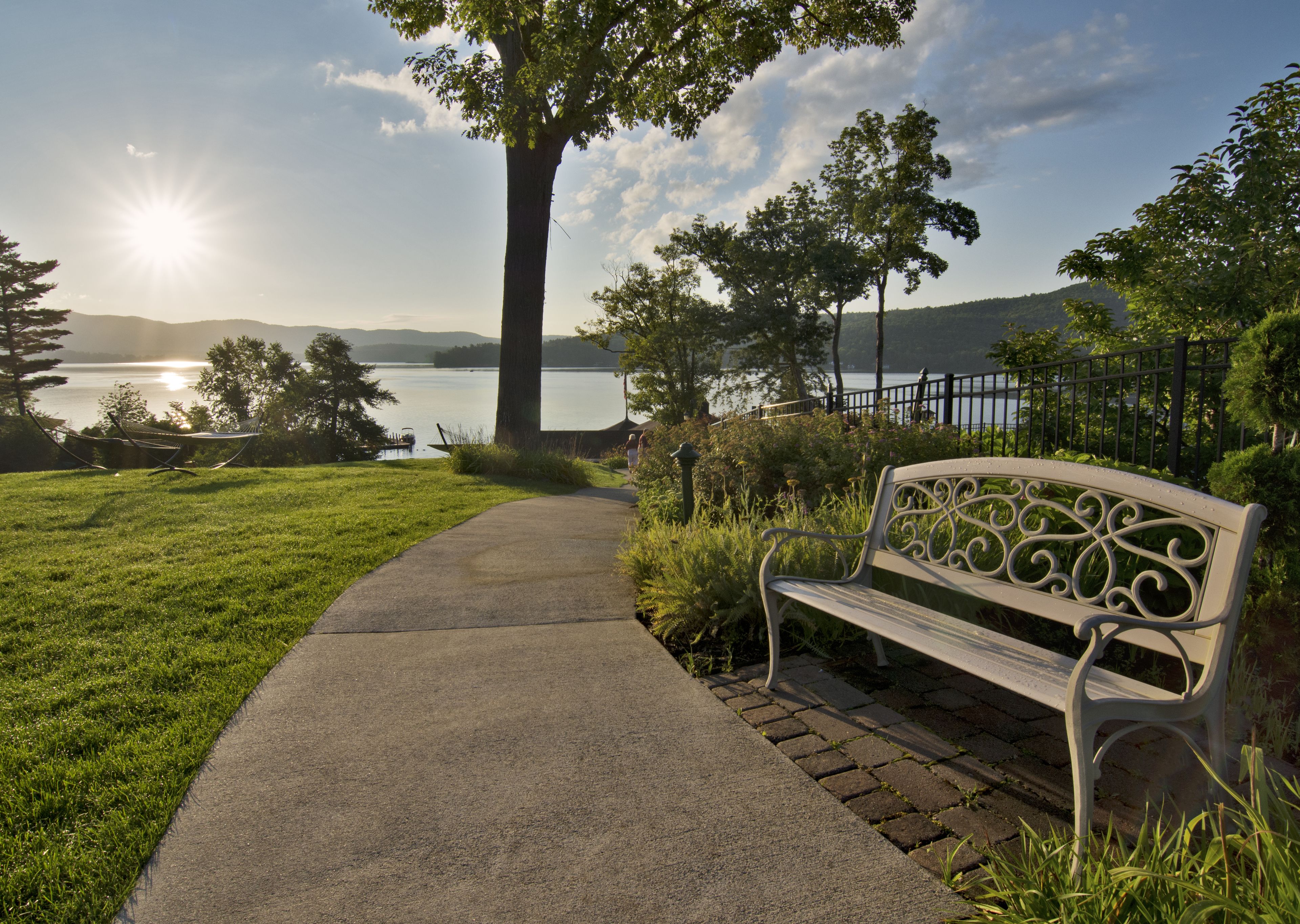 A park with a paved walkway, a bench, and a view of a lake with mountains in the background.
