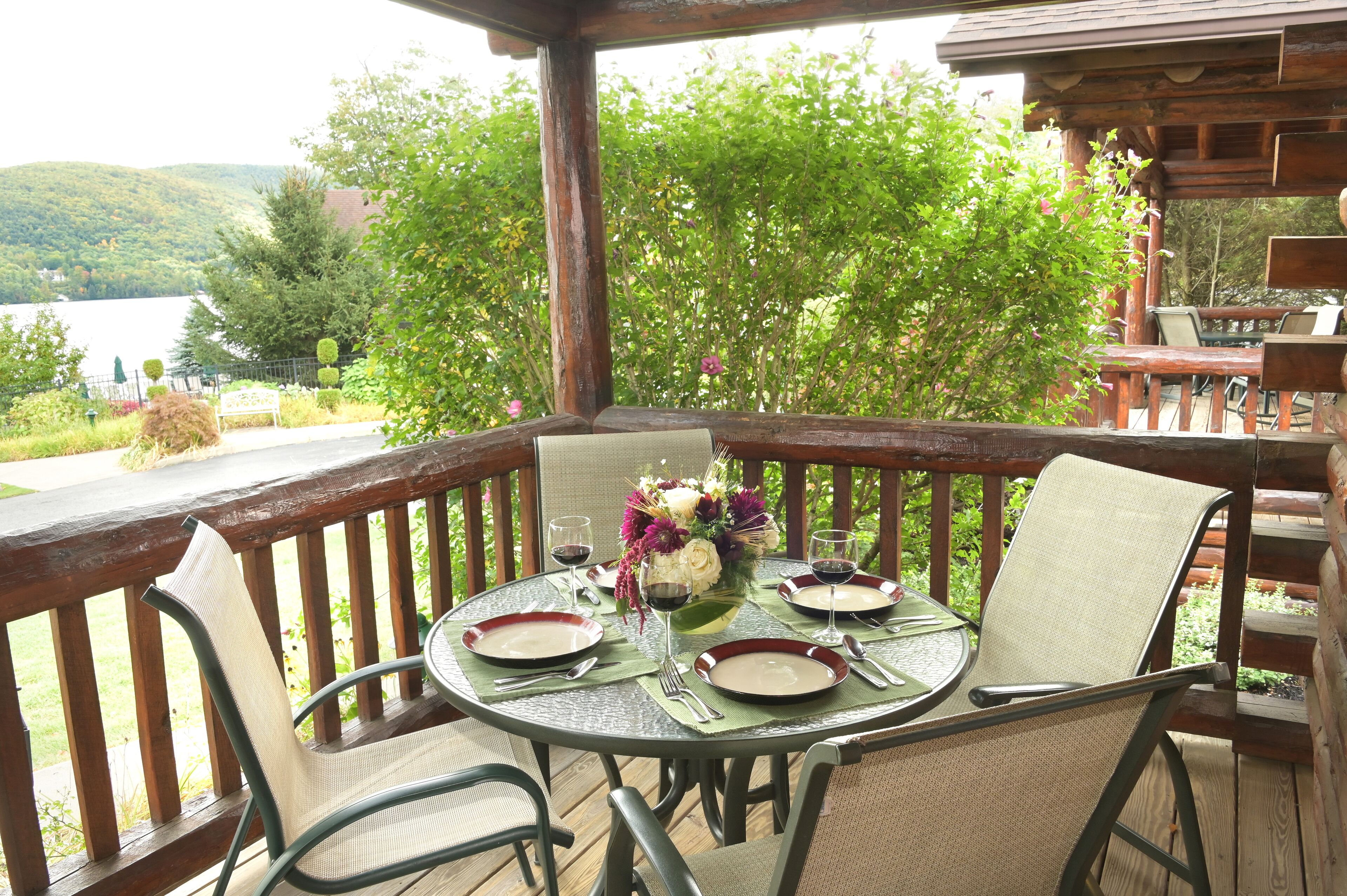 A wooden deck with a round table set for dining, overlooking a lake and surrounded by greenery.