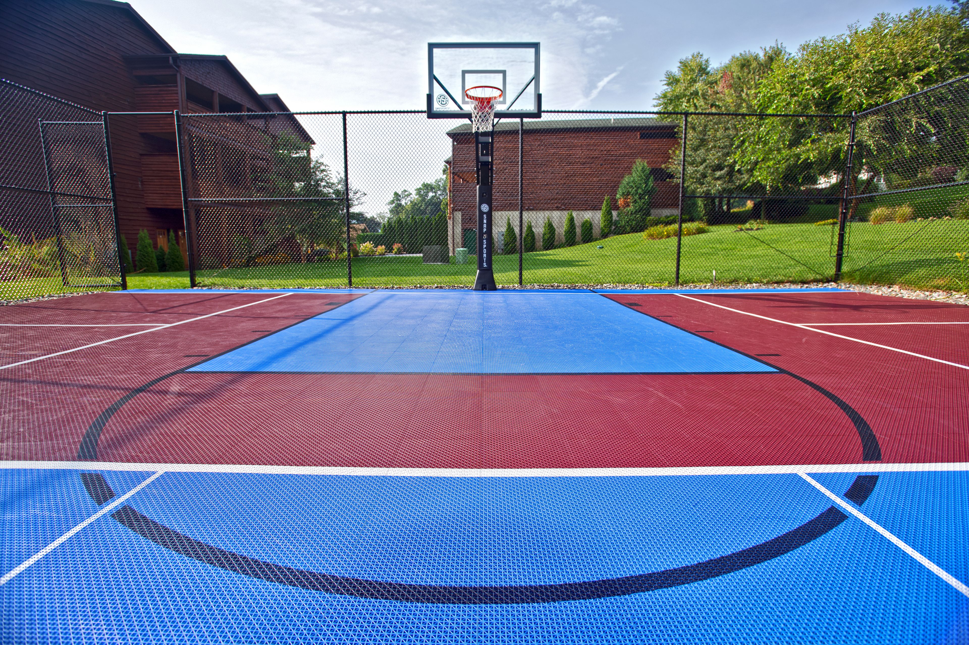 An outdoor basketball court with a red and blue playing surface, surrounded by a chain-link fence, and a building in the background.